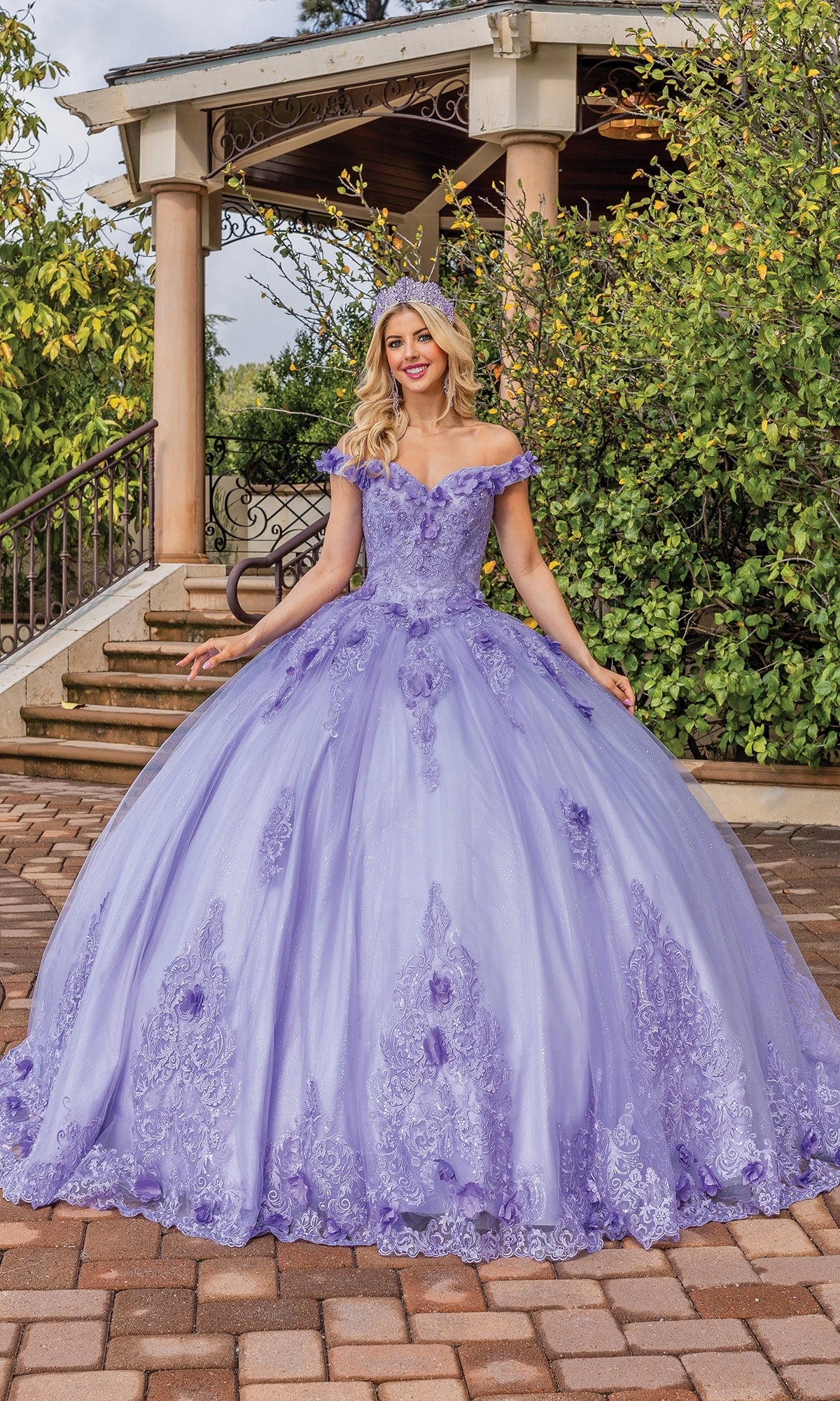 A woman smiles in front of a gazebo surrounded by greenery, wearing the Quinceanera Dress 1739 By Dancing Queen—an ornate, off-the-shoulder gown with delicate lace embroidery.
